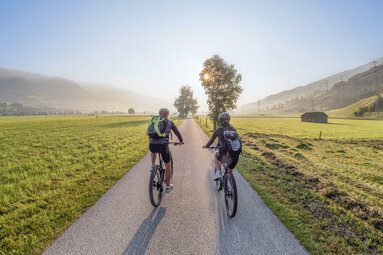Zwei Personen fahren auf dem Tauernradweg durch Wiesen bei Piesendorf und Niedernsill, im Hintergrund liegen die Berge. | © Ferienregion Nationalpark Hohe Tauern, Agentur Lichtfarben