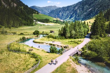 Zwei Personen mit Fahrrädern auf einem Weg im Talschluss Hüttschlag, neben einem Bach und umgeben von Bergen und Wald. | © Ferienregion Nationalpark Hohe Tauern, Foto Daniel Kogler