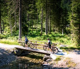 Zwei Personen mit Mountainbikes überqueren bei Hüttschlag am Ötzlsee eine Holzbrücke über einen Bach im Wald. | © Ferienregion Nationalpark Hohe Tauern, Foto Daniel Kogler