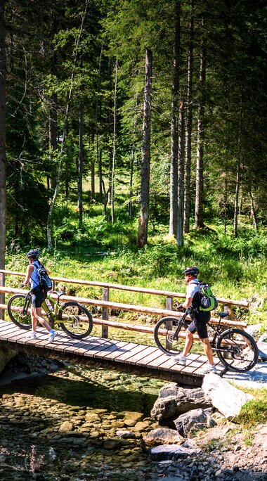 Zwei Personen mit Mountainbikes überqueren bei Hüttschlag am Ötzlsee eine Holzbrücke über einen Bach im Wald. | © Ferienregion Nationalpark Hohe Tauern, Foto Daniel Kogler
