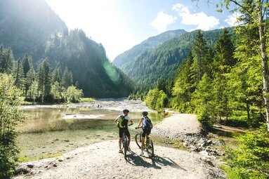 Zwei Personen mit Fahrrädern am Ötzlsee bei Hüttschlag, umgeben von bewaldeten Berghängen und einem breiten Bachbett. | © Ferienregion Nationalpark Hohe Tauern, Foto Daniel Kogler