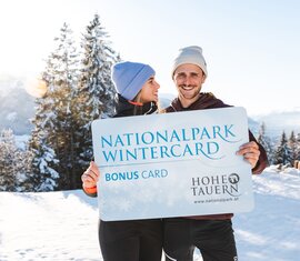 Two people hold the Nationalpark WinterCard in front of a snow-covered mountain landscape in Hohe Tauern. | © Ferienregion Nationalpark Hohe Tauern, Foto Mathäus Gartner 
