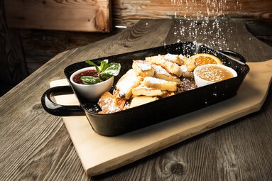 Kaiserschmarrn mit Puderzucker in einer schwarzen Pfanne, dazu zwei Beilagen und eine Orangenscheibe. | © SalzburgerLand Tourismus