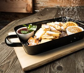 Kaiserschmarrn mit Puderzucker in einer schwarzen Pfanne, dazu zwei Beilagen und eine Orangenscheibe. | © SalzburgerLand Tourismus