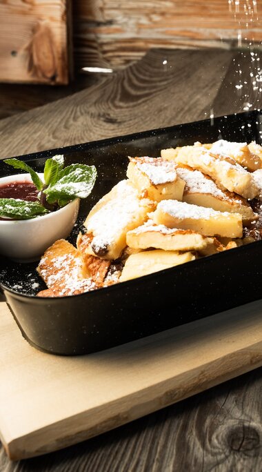 Kaiserschmarrn dusted with powdered sugar in a black pan, served with two side dishes and an orange slice. | © SalzburgerLand Tourismus