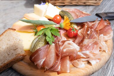 Jausenbrett mit Speck, Wurst, Käse, Brot, Essiggurken und Tomaten auf einem Holztisch. | © TVB Piesendorf Niedernsill, Foto Harry Liebmann 