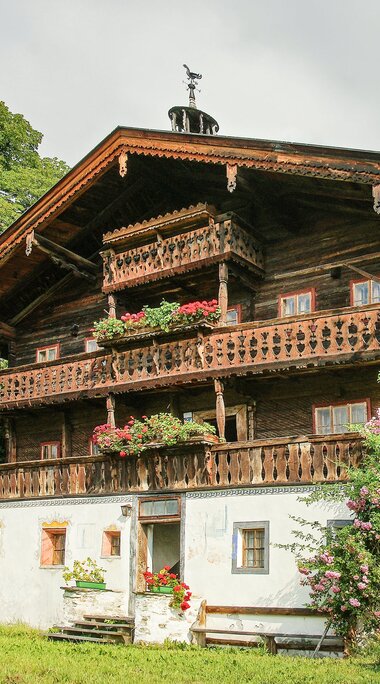 Ferchlergut in Fürth with a timber façade, decorated balconies and flowers, surrounded by meadows and trees. | © TVB Piesendorf Niedernsill, Foto Harry Liebmann 