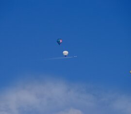 Three hot air balloons and a small plane with a contrail in the blue sky above clouds. | © TVB Piesendorf Niedernsill, Foto Harry Liebmann