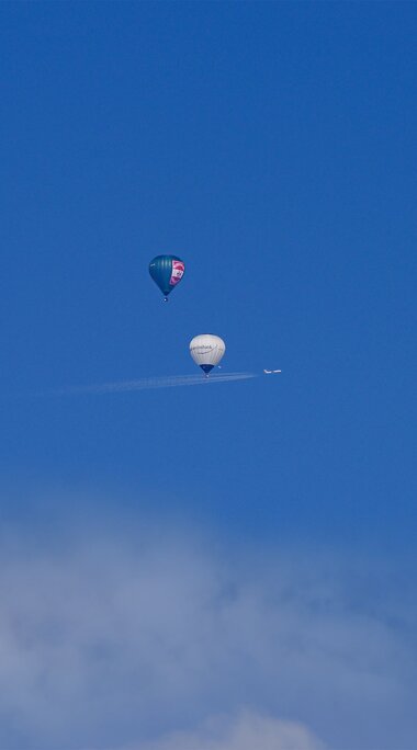 Three hot air balloons and a small plane with a contrail in the blue sky above clouds. | © TVB Piesendorf Niedernsill, Foto Harry Liebmann