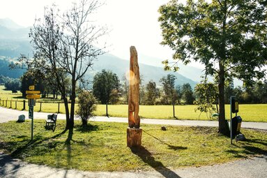 Holzskulptur „Kopfsteher“ von Sebastian Kusenberg auf einer Wiese zwischen Bäumen und Wegen, mit Bergen im Hintergrund. | © TVB Piesendorf Niedernsill, Foto Harry Liebmann