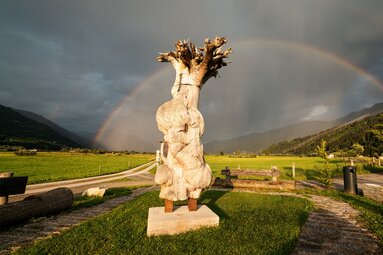 Holzskulptur zur Piesendorfer Grundzusammenlegung auf einer Wiese, dahinter ein Regenbogen über dem Tal und den Bergen. | © TVB Piesendorf Niedernsill, Foto Harry Liebmann