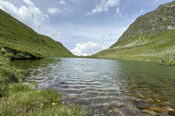 Schwarzkarlsee bei Uttendorf zwischen grünen, felsigen Berghängen, mit steinigem Ufer und Gräsern im Vordergrund. | © Julia Schuchter
