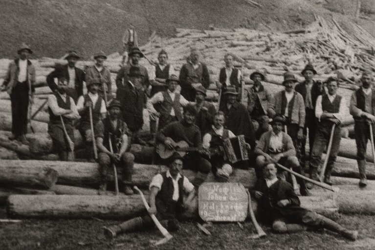 Holzknechte beim Schrempfbauer 1928 zwischen Baumstämmen, mit Werkzeugen, Gitarre und Ziehharmonika. | © TVB Piesendorf Niedernsill