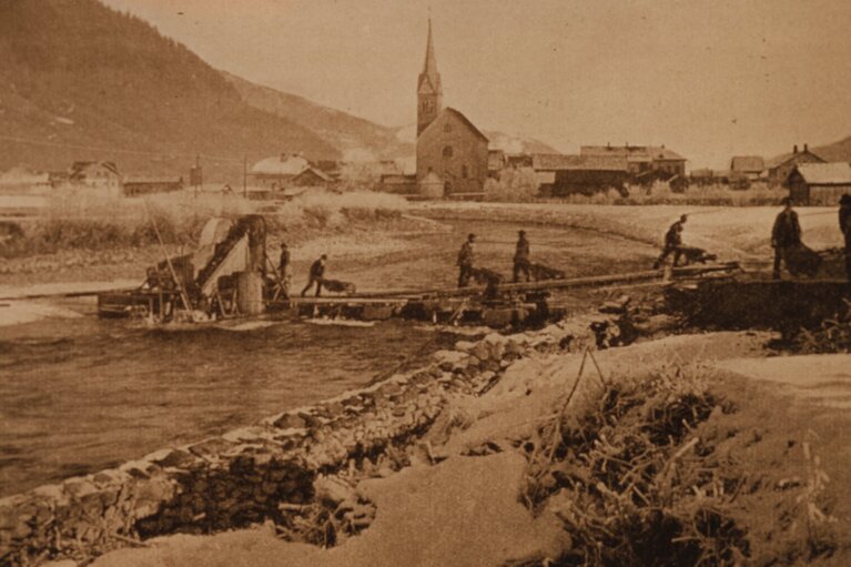 Historische Aufnahme der Salzachregulierung in Niedernsill mit Arbeitern am Gewässer und der Kirche im Hintergrund. | © TVB Piesendorf Niedernsill