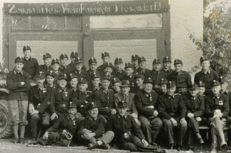 Historische Gruppenaufnahme der Freiwilligen Feuerwehr Piesendorf in Uniform vor ihrer Zeugstätte, links ein Traktor. | © TVB Piesendorf Niedernsill
