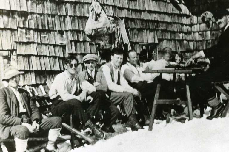 Historische Aufnahme der Piesendorfer Skipioniere vor einer Holzhütte, mit Skiern im Vordergrund. | © TVB Piesendorf Niedernsill