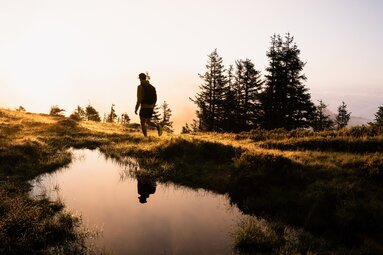 Trail runner on stage 09 of the Hohe Tauern Panorama Trail in low sunlight beside a small body of water. | © Ferienregion Nationalpark Hohe Tauern, Gerald Demolsky