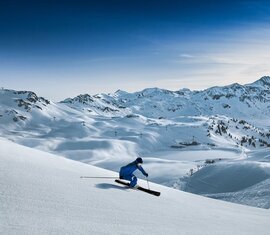 Skier on a snow-covered slope with a wide mountain panorama and ski lifts in the background. | © Rohrbacher