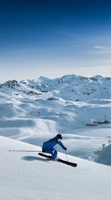 Skier on a snow-covered slope with a wide mountain panorama and ski lifts in the background. | © Rohrbacher