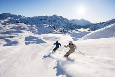 Two people ski downhill on a groomed slope with snow-covered mountains in the background. | © SuperSkiCard | Mr-Offenblende.at