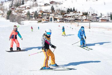 Children ski and snowboard at Erlebnisberg Naglköpfl in Piesendorf, with houses on the snowy hillside in the background. | © TVB Piesendorf Niedernsill, Foto Harry Liebmann 