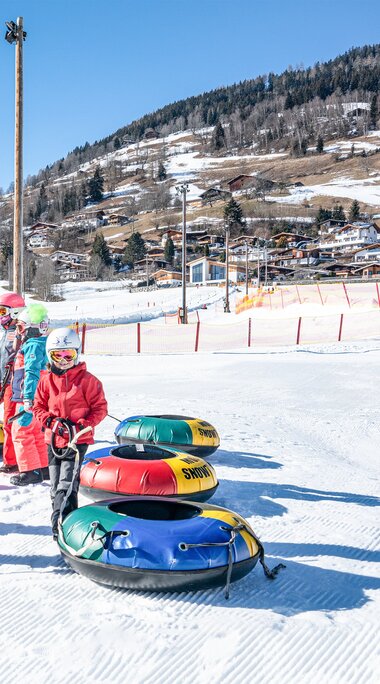 Kinder mit Helmen stehen mit bunten Snowtubes auf der Schneefläche am Erlebnisberg vor einem verschneiten Hang. | © TVB Piesendorf Niedernsill, Foto Harry Liebmann