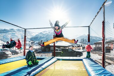 Children jump on a net-enclosed trampoline area at Erlebnisberg Naglköpfl with snow-covered mountains behind them. | © TVB Piesendorf Niedernsill, Foto Harry Liebmann