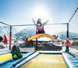 Children jump on a net-enclosed trampoline area at Erlebnisberg Naglköpfl with snow-covered mountains behind them. | © TVB Piesendorf Niedernsill, Foto Harry Liebmann
