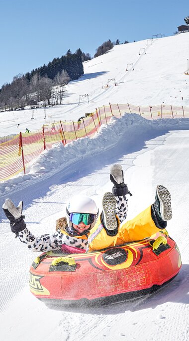 Kind mit Helm fährt in einem roten Snowtube eine präparierte, mit Fangnetzen begrenzte Schneebahn am Erlebnisberg hinunter. | © TVB Piesendorf Niedernsill, Foto Harry Liebmann