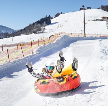 Kind mit Helm fährt in einem roten Snowtube eine präparierte, mit Fangnetzen begrenzte Schneebahn am Erlebnisberg hinunter. | © TVB Piesendorf Niedernsill, Foto Harry Liebmann