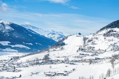 Snow-covered valley with villages, a ski slope and surrounding mountains under a blue sky. | © TVB Piesendorf Niedernsill