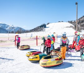 Children wearing helmets stand with colourful snow tubes at Erlebnisberg, with a snow-covered slope in the background. | © TVB Piesendorf Niedernsill, Foto Harry Liebmann