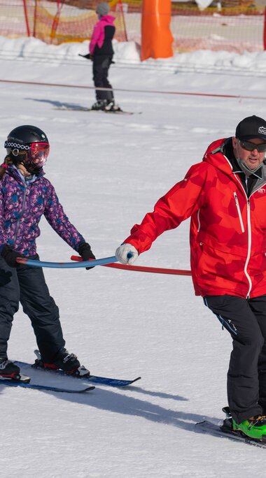 Ski instructor from Skischule Entleitner assists a child during a skiing exercise using a training bar. | © Florian Entleitner, Schischule Entleitner