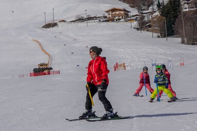 Ski instructor leads three children practising with coloured poles on a groomed ski slope. | © Florian Entleitner, Schischule Entleitner