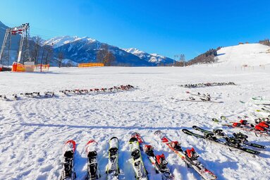 Numerous skis lie in the snow beside the slope at Erlebnisberg Naglköpfl in Piesendorf, with mountains in the background. | © Lichtfarben