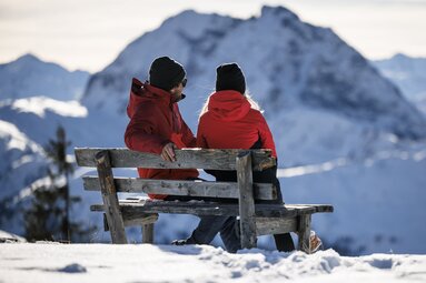 Two people sitting on a wooden bench overlooking snow-covered mountains in the Kitzbühel Alps. | © Mediahouse, Max Draeger