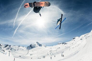 Snowboarder and skier jump simultaneously in the snowpark at Kitzsteinhorn against snow-covered mountains. | © Kitzsteinhorn