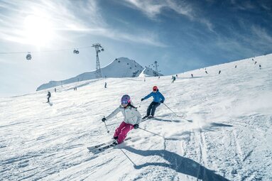 Zwei Personen fahren am Kitzsteinhorn Ski. Im Hintergrund sind weitere Skifahrer sowie Gondeln zu sehen. | © Kitzsteinhorn