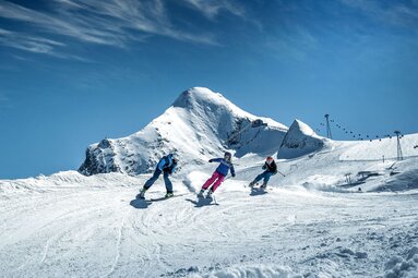 Three people ski along the Funline at Kitzsteinhorn, with a snow-covered peak and ski lifts in the background. | © Kitzsteinhorn