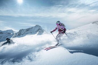 Two people skiing through deep snow with snow-covered mountains in the background. | © Kitzsteinhorn