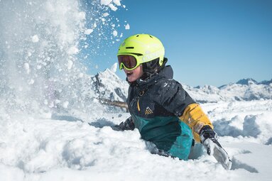 Kind mit Skihelm spielt im Schnee im Skicircus Saalbach Hinterglemm Leogang Fieberbrunn, dahinter verschneite Berge. | © saalbach.com, Klaus Listl