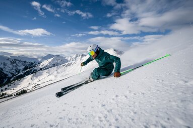 Skifahrer fährt eine Kurve im Skicircus Saalbach Hinterglemm Leogang Fieberbrunn, dahinter erstrecken sich verschneite Berge. | © saalbach.com, Stefan Voitl