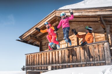 Zwei Kinder in Skibekleidung springen vor einer verschneiten Holzhütte in die Luft, zwei Erwachsene stehen hinter ihnen. | © saalbach.com, Klaus Listl