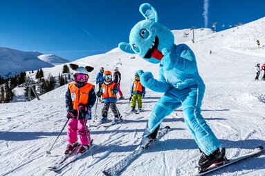Maskottchen Slopy fährt mit mehreren Kindern Ski auf einer verschneiten Piste, im Hintergrund sind Liftanlagen zu sehen. | © Christian Riefenberg