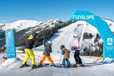 Zwei Erwachsene und zwei Kinder auf Ski am gekennzeichneten Eingang der Funslope Schmitten. | © Schmittenhöhebahn AG, Foto Michael Schröder