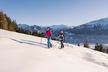 Two women hike on snowshoes at Plettsaukopf with snow-covered mountains in the background. | © Schmittenhöhebahn AG