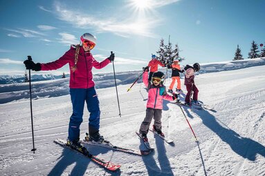 Erwachsene und Kinder stehen gemeinsam mit Ski auf einer sonnigen, präparierten Piste auf der Schmittenhöhe. | © Schmittenhöhebahn AG