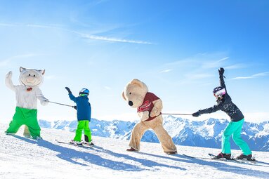 Two skiers with two mascots at Kogel-Mogel Kinderland in Wildkogel-Arena, with snow-covered mountains behind them. | © Wildkogel-Arena Neukirchen & Bramberg