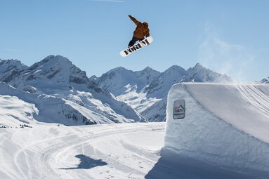 Snowboarder jumps over a snow feature at Action Park Kreuzwiese in Zillertal Arena, with snow-covered mountains behind. | © Zillertal Arena