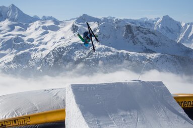 Freestyle skier jumps over a snow ramp at Action Park Kreuzwiese in Zillertal Arena, with snow-covered mountains behind. | © Zillertal Arena